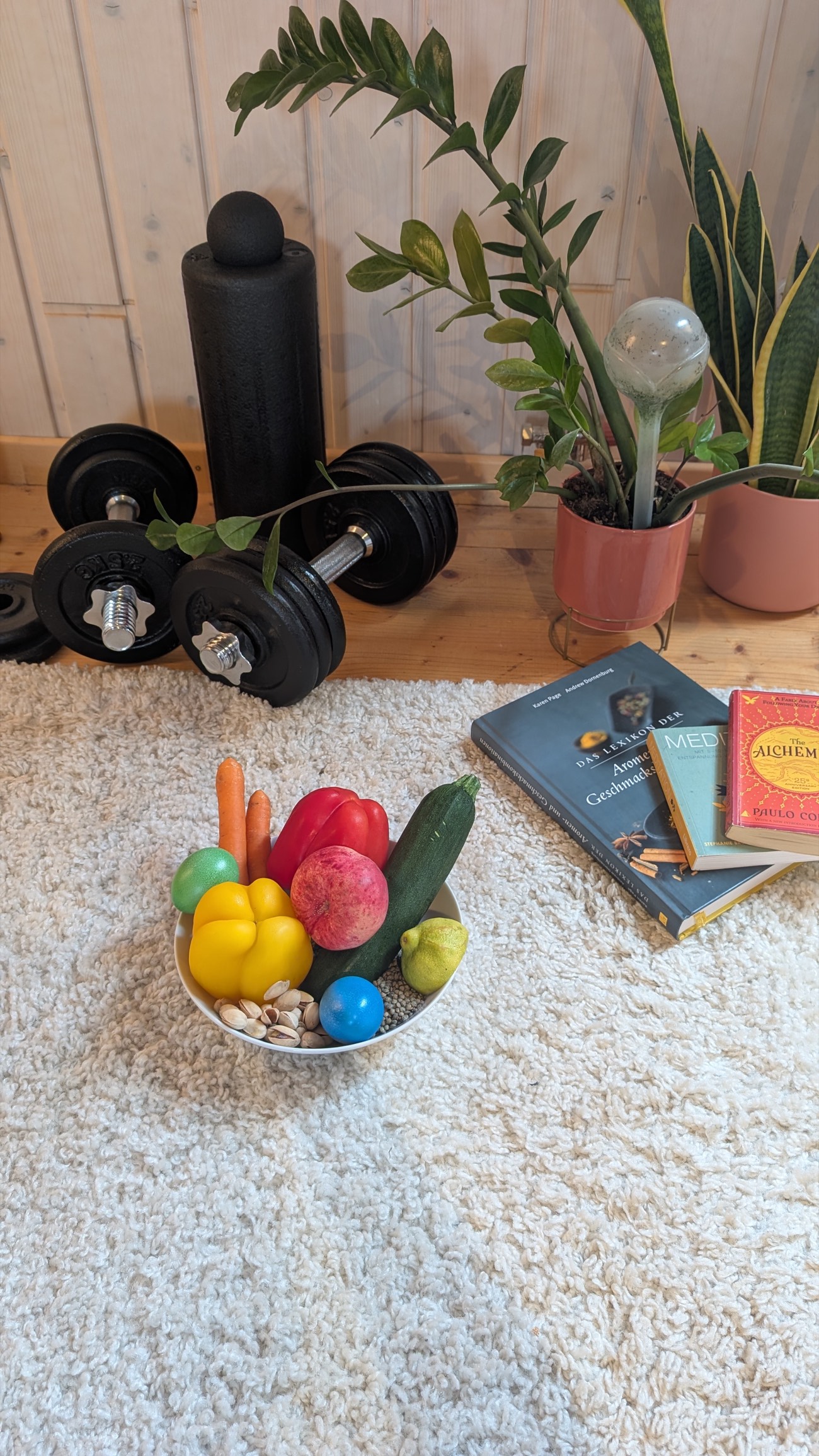 Schale mit frischem Obst und Gemüse auf dem Teppich, daneben Bücher und Hanteln